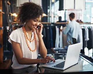 A woman sitting at the desk of her clothing boutique and talking on her phone while looking at her laptop